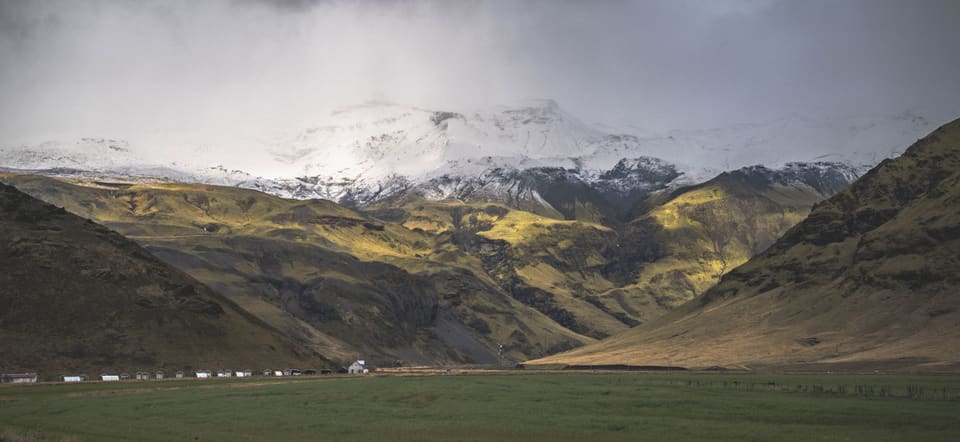 A green valley under a snow-covered volcano in south Iceland, with a row of small white farm buildings along its floor and storm cloud above.
